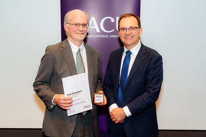  Man with glasses and grey hair holding certificate beside another man in dark suit wearing glasses