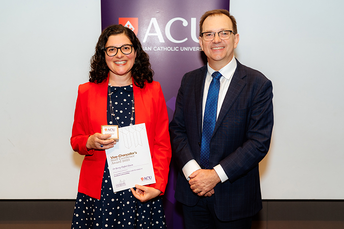  Woman with dark hair and glasses in red jacked holding a certificate and plaque beside man in dark suit and glasses