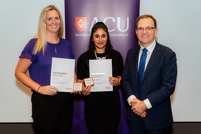  Woman with long blonde hair in purple top holding certificate with woman with dark hair and black top holding certificate beside man with glasses in dark suit