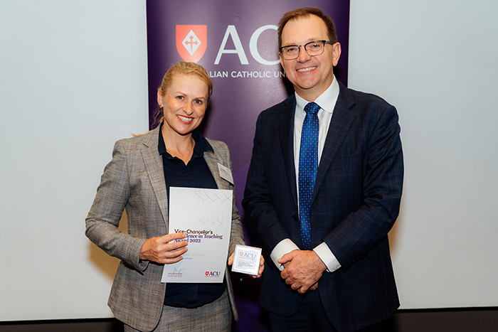  Woman with blonde hair in ponytail holding a certificate beside a man in glasses and dark suit
