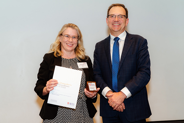  Woman with blonde shoulder length hair and glasses in black and white dress holds a certificate and plaque with a man in a dark suit and glasses beside her