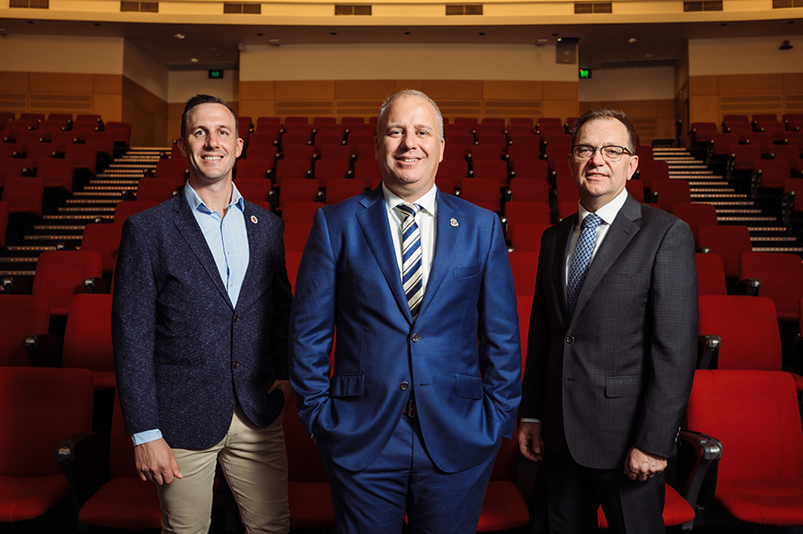  Three men - Aaron Cornwall a white man in a dark blue blazer and blue shirt, RSL Queensland CEO Robert Skoda a white man in a blue suit, and Professor Zlatko Skrbis a white man in a dark grey suit - stand in a lecture theatre