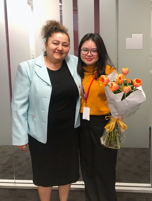  a woman in a black dress and pale blazer with a woman in an orange turtleneck and black trousers holding a bunch of tulips